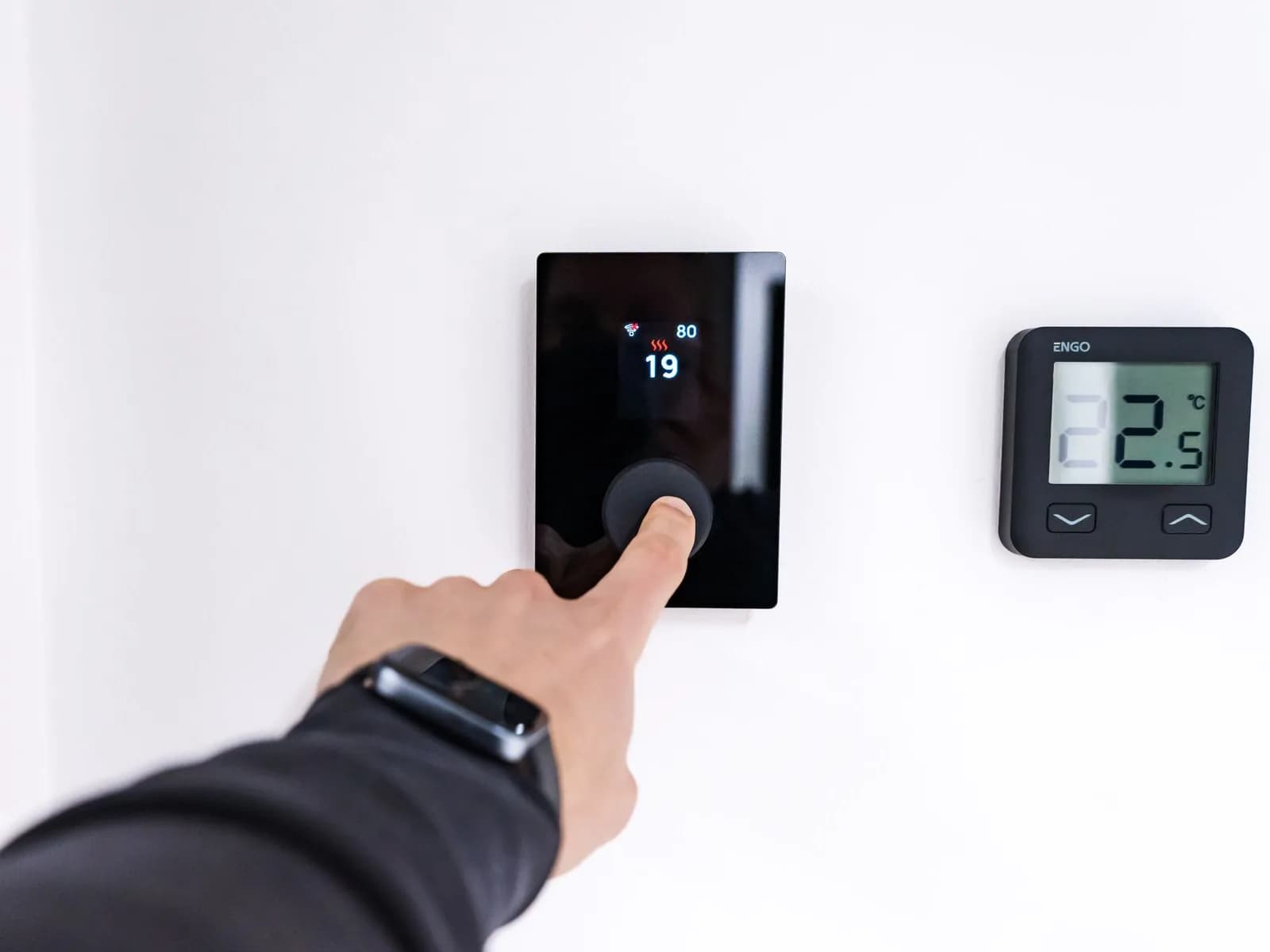 A hand using a wall-mounted touchscreen beside a digital thermostat in a modern home.